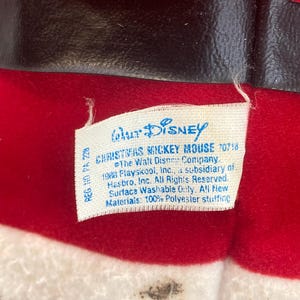 May include: A red plush fabric toy with a black belt and white trim, featuring a white tag that reads Walt Disney Christmas Mickey Mouse 70718, The Walt Disney Company, 1988 Playskool, Inc., a subsidiary of Hasbro, Inc. All Rights Reserved.
