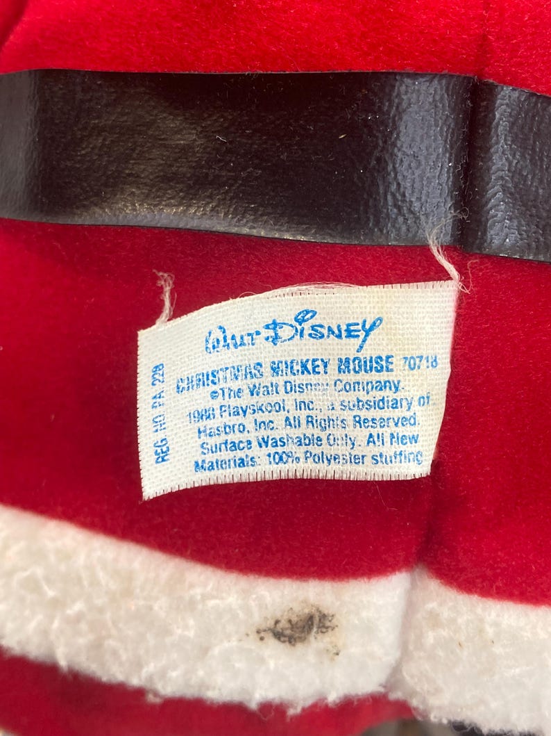 May include: A red plush fabric toy with a black belt and white trim, featuring a white tag that reads Walt Disney Christmas Mickey Mouse 70718, The Walt Disney Company, 1988 Playskool, Inc., a subsidiary of Hasbro, Inc. All Rights Reserved.