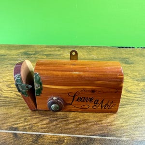 May include: A wooden box with a hinged lid and a small knob. The box is a warm brown color with the words "Leave a Note" in cursive script. A metal clasp and a small metal loop are visible.