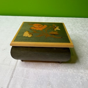 May include: A wooden music box with a green top featuring a floral design in shades of orange and yellow. The box has a dark grey, curved body and rests on small feet. The top is framed with a light wood border.
