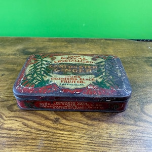 May include: A rectangular, antique tin with a red and black exterior. The tin features text that reads "LYONS ROYAL CRYSTALLIZED CANTON STEM GINGER" and "CALIFORNIA GLACE FRUIT CO." in white and green lettering. The tin is on a wooden surface.