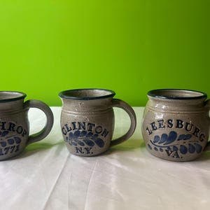 May include: Three stoneware mugs with blue and white designs. The mugs feature the names of towns in the United States: Winthrop, Est. 1771, Clinton, NY, and Leesburg, VA.