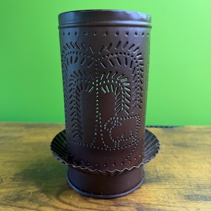 May include: A cylindrical brown metal candle holder featuring a punched tin design of a weeping willow tree and a small sheep. The metal surface has a matte finish with a scalloped edge base, creating a decorative pattern through the perforations.