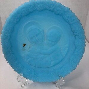 May include: A light blue glass plate with a raised design of two figures holding hands. The plate has a white border with a floral design.