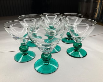 May include: Set of six clear glass stemmed drinking glasses with green bases. The glasses are arranged in a circle on a white surface.
