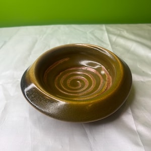 May include: A handcrafted ceramic bowl featuring a spiral pattern at its centre. The bowl has a rounded form, with a dark green exterior and a lighter green interior. The spiral design is reddish-brown.