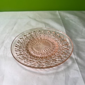 Op de afbeelding: Een heldere, roze glazen plaat met een ruitpatroon rond de rand en een sterrenpatroon in het midden. De plaat is cirkelvormig en ligt op een wit oppervlak, met een groene achtergrond.