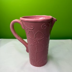 Op de afbeelding: Een roze keramische kan met een handvat en een tuit. De kan is versierd met een reliëf bloemenontwerp. De achtergrond is effen groen.
