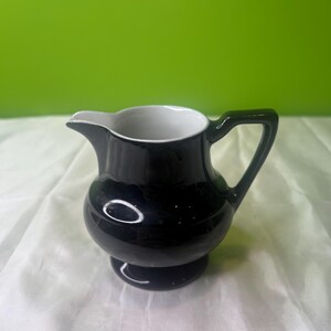 May include: A small black creamer with a white interior and a rounded body. The creamer has a handle and a small spout.