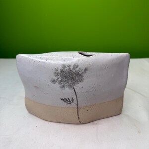 May include: A handmade ceramic vase with a light gray upper section and a tan base. A black and gray floral design is painted on the front. The vase has a unique, asymmetrical shape and a small opening at the top.