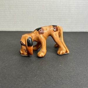 May include: A small ceramic figurine of a brown and black dog with its head down, as if sniffing the ground. The dog has brown and black spots and is standing on all four legs.