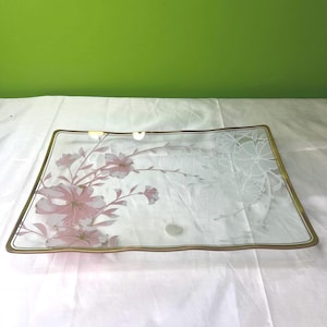 Puede incluir: Bandeja rectangular de vidrio con borde dorado. La bandeja presenta un diseño floral en rosa y blanco, con un aspecto delicado y grabado. Las flores y hojas están dispuestas sobre la superficie, creando una pieza elegante y decorativa.