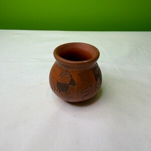 May include: A small, round, terracotta-colored pot with a wide opening. The pot features etched black designs of animals and abstract shapes around its exterior. The background is a solid green color.