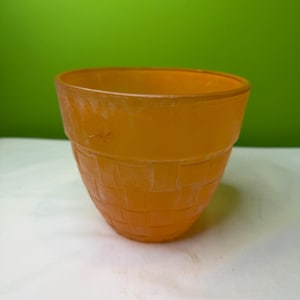 Pode incluir: Um vaso de flores laranja, texturizado e em forma de cone. O vaso tem um padrão de tecido de cesta no exterior. O vaso é feito de um material translúcido e está vazio. O fundo é de cor verde lisa.