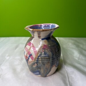 May include: A small, handcrafted ceramic vase with a round body and flared rim. The vase features a cream-colored base with blue, pink, and brown abstract patterns. The interior rim is a deep blue. The vase is approximately 5 inches tall.