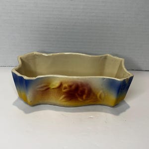 May include: A small, white ceramic planter with a blue and yellow glaze. The planter has a scalloped edge and a floral design in brown.