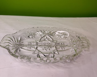 May include: Clear glass serving dish with three compartments and starburst and geometric designs. The oval-shaped dish has handles on each end. The glass is transparent, allowing light to pass through, and is suitable for serving appetizers or snacks.