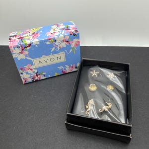 May include: A set of six gold-tone metal pins in a black box, featuring two starfish, two seashells, and two seahorses. The pins are covered by a clear plastic film. The box lid is light blue with a floral pattern and the text AVON printed on it.