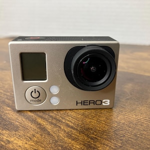 May include: A silver HERO3 action camera with a black lens and a small LCD screen. The camera has a power button and a mode button. The camera is on a wooden surface.