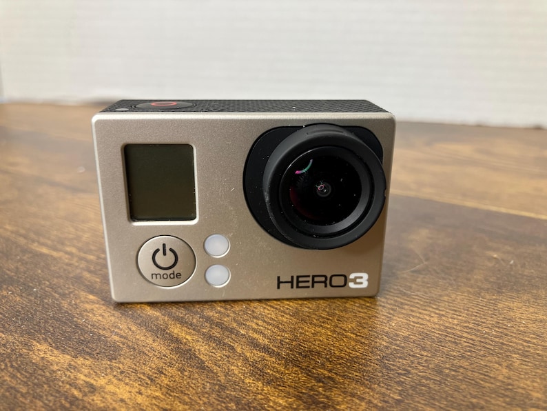 May include: A silver HERO3 action camera with a black lens and a small LCD screen. The camera has a power button and a mode button. The camera is on a wooden surface.