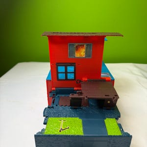 May include: A red toy train caboose with a brown roof and a small brown wooden deck. The caboose has a window with a fire graphic. The base is blue and green with a small scooter graphic.