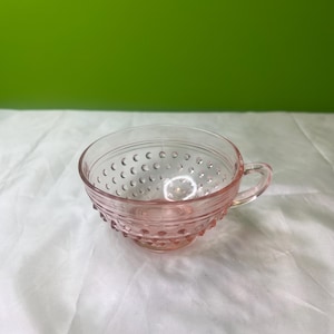 以下が含まれることがあります： ピンク色のガラスのティーカップ。取っ手が付いています。カップは、ボウルとベースの周りに小さな隆起したドットのテクスチャード加工が施されています。透明なカップは光を通します。