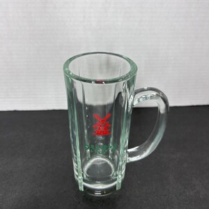 May include: A clear glass shot glass with a handle. The glass has a red windmill logo with green lettering that says "Prins."