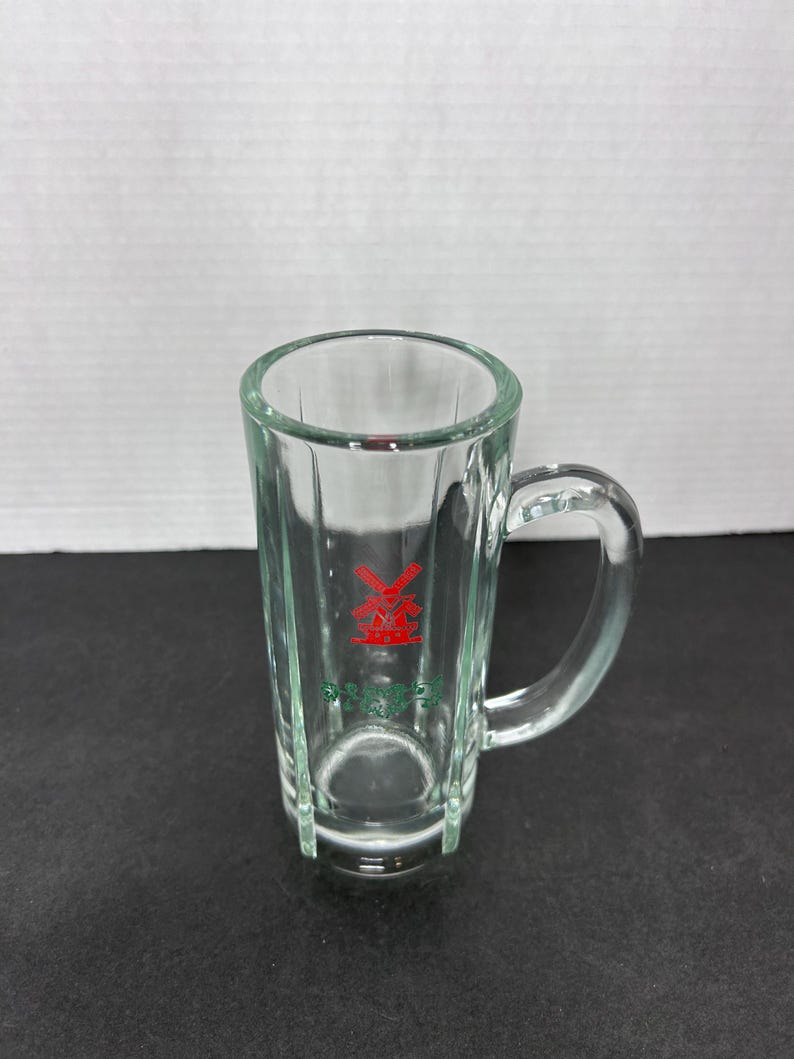 May include: A clear glass shot glass with a handle. The glass has a red windmill logo with green lettering that says "Prins."