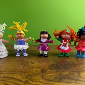 May include: Five Cabbage Patch Kids figurines in various outfits. The dolls have yarn hair and are holding small accessories. The dolls are on a wooden surface with a green background.