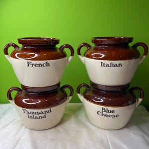 Puede incluir: Cuatro cuencos de cerámica para servir con bordes y asas marrones, apilados en pares. Los cuencos son blancos con detalles marrones y presentan las palabras "French", "Italian", "Thousand Island" y "Blue Cheese" en texto negro.