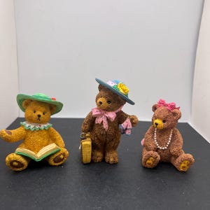 Peut inclure: Trois figurines décoratives d'ours. L'un est jaune et lit un livre, portant un chapeau vert. Le second est marron, tenant une valise et portant un chapeau bleu. Le troisième est marron, avec un nœud rose et un collier de perles.