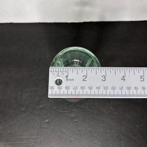 May include: A clear glass shot glass with a rounded bottom and a lip around the top. The shot glass is 1 inch in diameter.