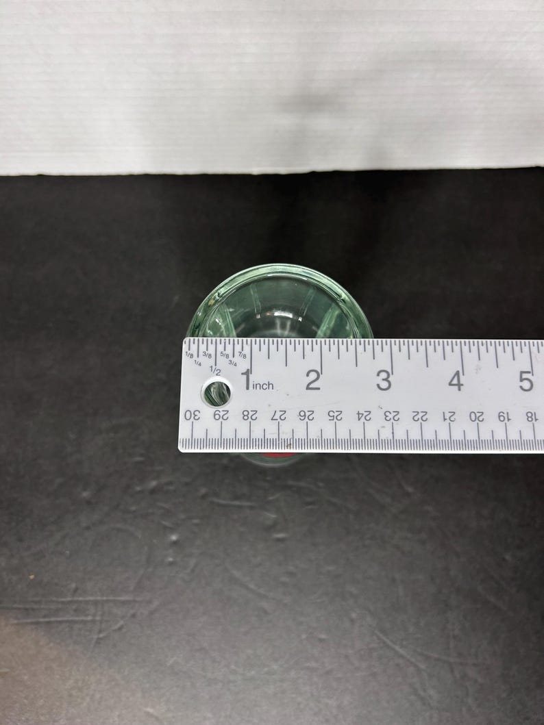 May include: A clear glass shot glass with a rounded bottom and a lip around the top. The shot glass is 1 inch in diameter.