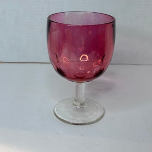 Pode incluir: Um copo de vinho de vidro transparente com uma taça de cor vermelha escura, quase bordô. A taça tem uma superfície texturizada, ligeiramente irregular.