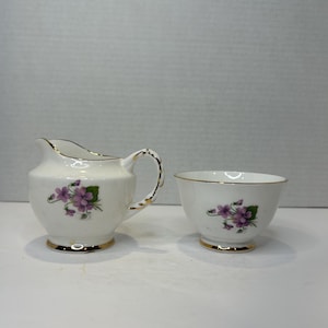 Puede incluir: Un juego de jarra de crema y azucarero blanco con un diseño floral con flores moradas y hojas verdes. El juego tiene un borde dorado.