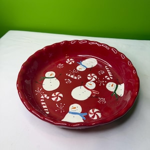 May include: A red ceramic pie dish with a scalloped edge. The dish is decorated with white snowmen wearing scarves and surrounded by candy canes and snowflakes. A festive holiday design.