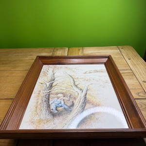 Könnte beinhalten: Ein gerahmter Druck mit einem Kind in blauen Latzhosen, das in der Nähe einer Holzkonstruktion sitzt, umgeben von Bäumen. Das Kunstwerk hat eine weiche, gedämpfte Farbpalette mit Braun-, Beige- und Grüntönen. Der Rahmen ist aus dunklem Holz.