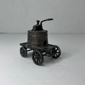 May include: A vintage metal pencil sharpener shaped like a horse-drawn cart with a barrel on top. The sharpener is painted in a dark brown color and has a handle on the side.
