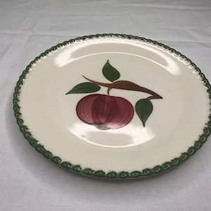 May include: A white ceramic dinner plate with a green scalloped edge. The plate features a hand-painted design of a red apple on a green branch with leaves.