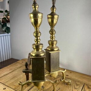 May include: Two antique brass fireplace andirons. The andirons have a tall, ornate design with a gold finish. Each andiron has a square base with a decorative foot and a tall, slender body. The andirons are displayed on a wooden table.