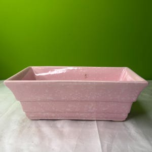 May include: A rectangular, light pink planter with a speckled texture. The planter has a stepped design and a slightly flared rim. The background is a solid green color.