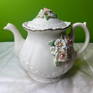 May include: Cream-coloured ceramic teapot with floral embellishments. The teapot has a lace trim around the base and lid, and is decorated with sculpted flowers in pink, white, and green. The handle and spout are curved.