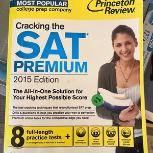 Puede incluir: Un libro amarillo y azul de preparación para el SAT titulado "Cracking the SAT Premium 2015 Edition" de The Princeton Review. La portada presenta a una mujer sonriente sosteniendo libros e información sobre pruebas.