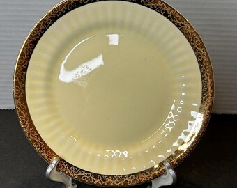 Puede incluir: Un plato de cerámica blanco con un borde dorado y un diseño floral dorado. El plato tiene un borde festoneado y está sobre un soporte blanco.