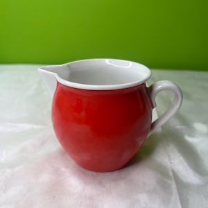 Może przedstawiać: Mały, czerwony ceramiczny dzbanek z białym wnętrzem i uchwytem. Dzbanek ma zaokrąglony korpus i dzióbek do nalewania. Uchwyt jest zakrzywiony i przymocowany do boku dzbanka. Tło jest jednolicie zielone.