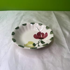 May include: A small, white ceramic bowl with a floral design. The bowl features a red flower with green leaves and a yellow flower. The rim is decorated with green, dotted patterns.