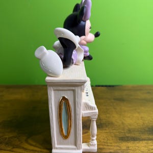 Peut inclure: Une figurine en c&eacute;ramique repr&eacute;sentant une souris noire et blanche avec un n&oelig;ud violet, allong&eacute;e sur un piano droit de couleur cr&egrave;me. Le piano pr&eacute;sente une texture imitation bois, un ovale dor&eacute; sur le c&ocirc;t&eacute; et un vase blanc sur le dessus.