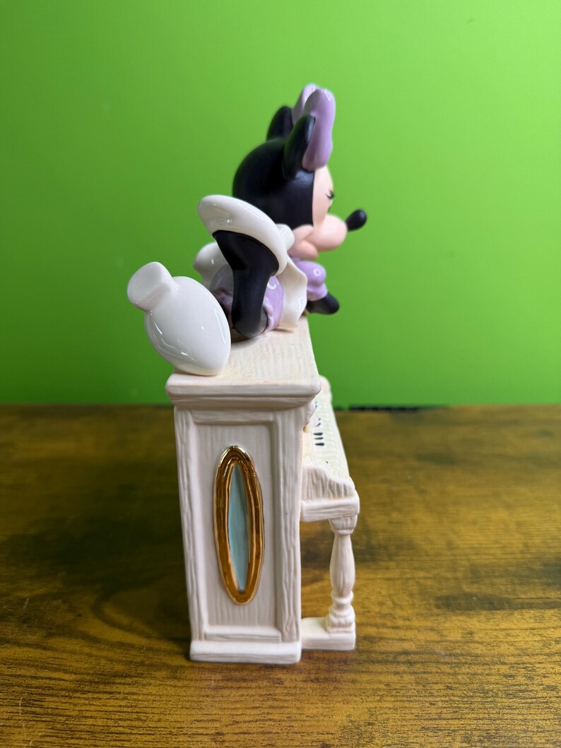 Peut inclure: Une figurine en c&eacute;ramique repr&eacute;sentant une souris noire et blanche avec un n&oelig;ud violet, allong&eacute;e sur un piano droit de couleur cr&egrave;me. Le piano pr&eacute;sente une texture imitation bois, un ovale dor&eacute; sur le c&ocirc;t&eacute; et un vase blanc sur le dessus.