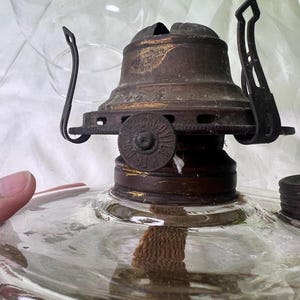 Puede incluir: Primer plano de una l&aacute;mpara de aceite antigua. La l&aacute;mpara tiene una base de vidrio transparente y una parte superior de metal oscuro con una mecha. La parte superior tiene un dise&ntilde;o decorativo y un asa. Una chimenea de vidrio transparente est&aacute; en el fondo.