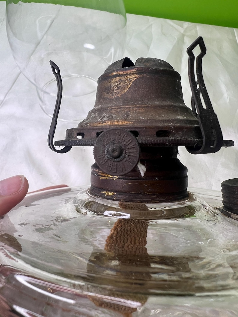 Puede incluir: Primer plano de una l&aacute;mpara de aceite antigua. La l&aacute;mpara tiene una base de vidrio transparente y una parte superior de metal oscuro con una mecha. La parte superior tiene un dise&ntilde;o decorativo y un asa. Una chimenea de vidrio transparente est&aacute; en el fondo.